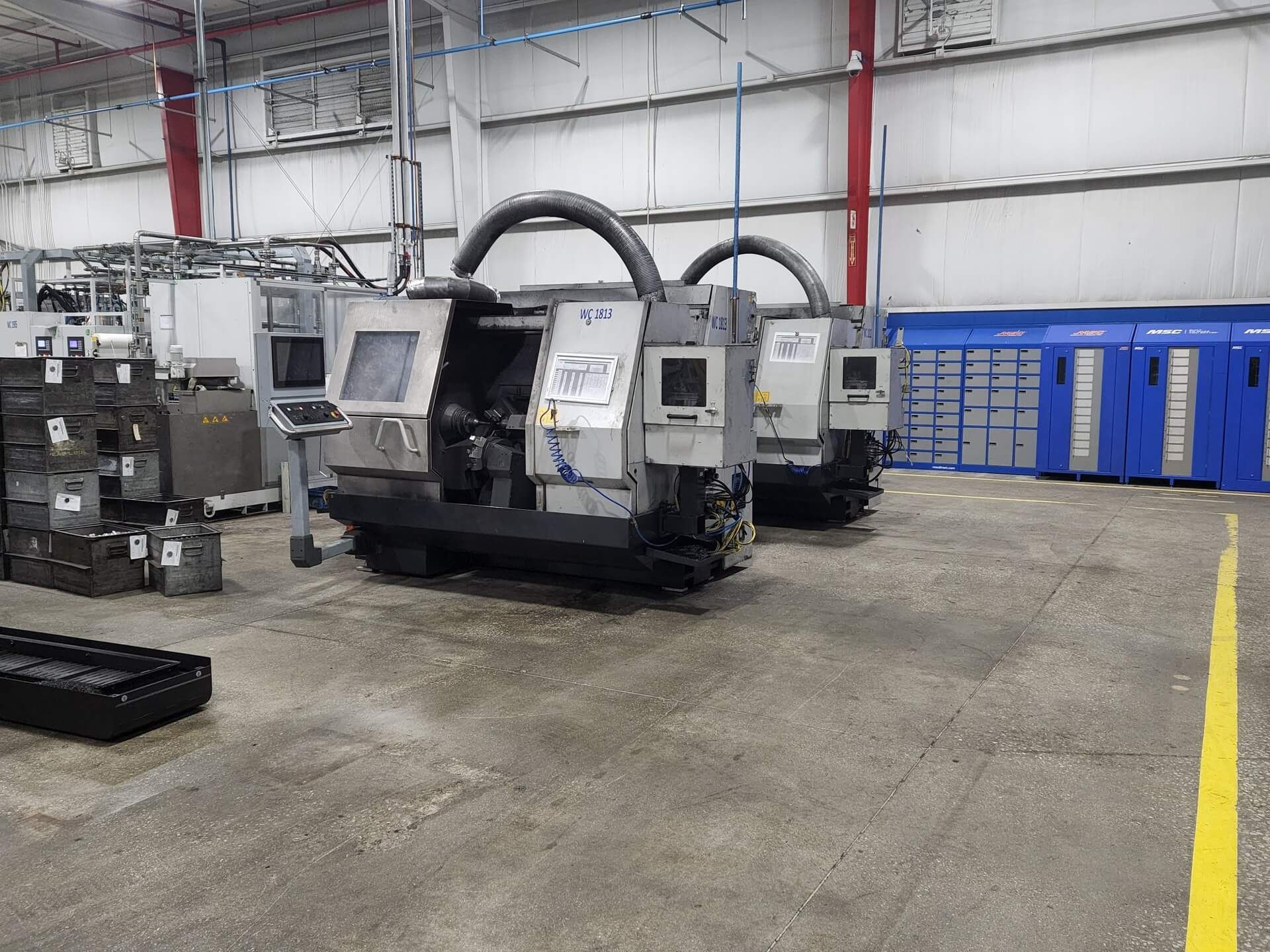 Two CNC machines in a factory setting. Grey machines with black tubing, concrete floor, and blue storage.
