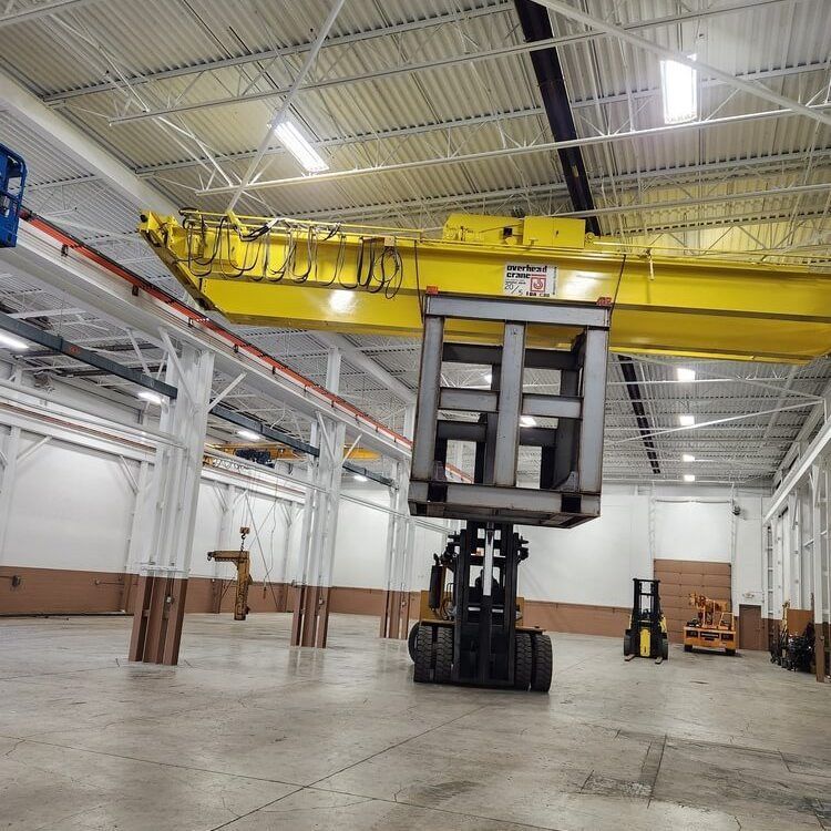 Forklift holding a large metal cage in a warehouse with a yellow overhead crane.