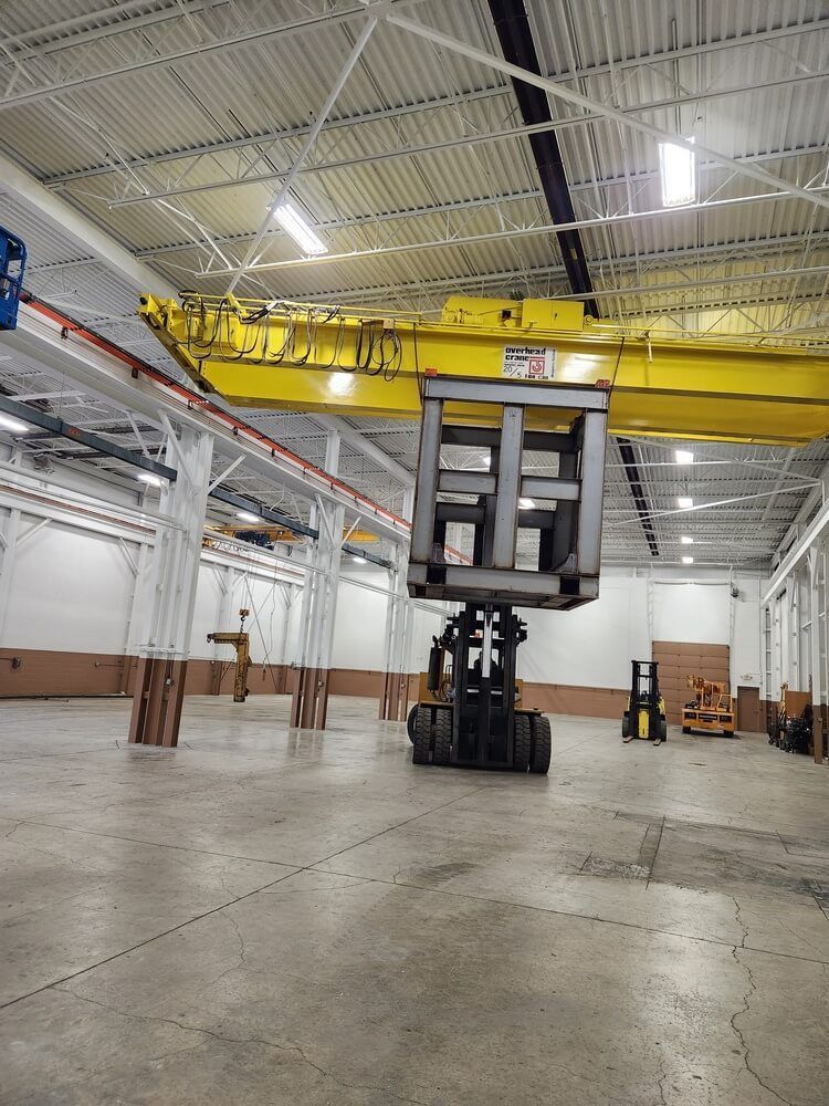Forklift lifting a large metal frame in a warehouse with a yellow overhead crane.