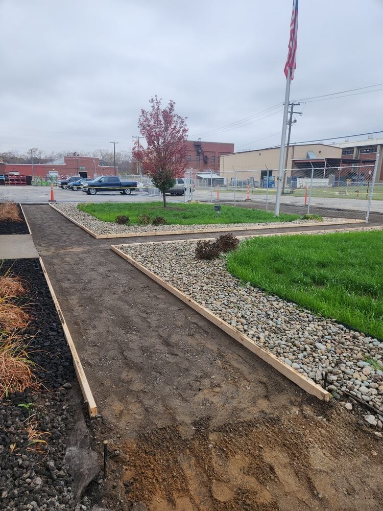 Pathway under construction, lined with wood, beside a rock garden and grass, with a red-leaved tree.
