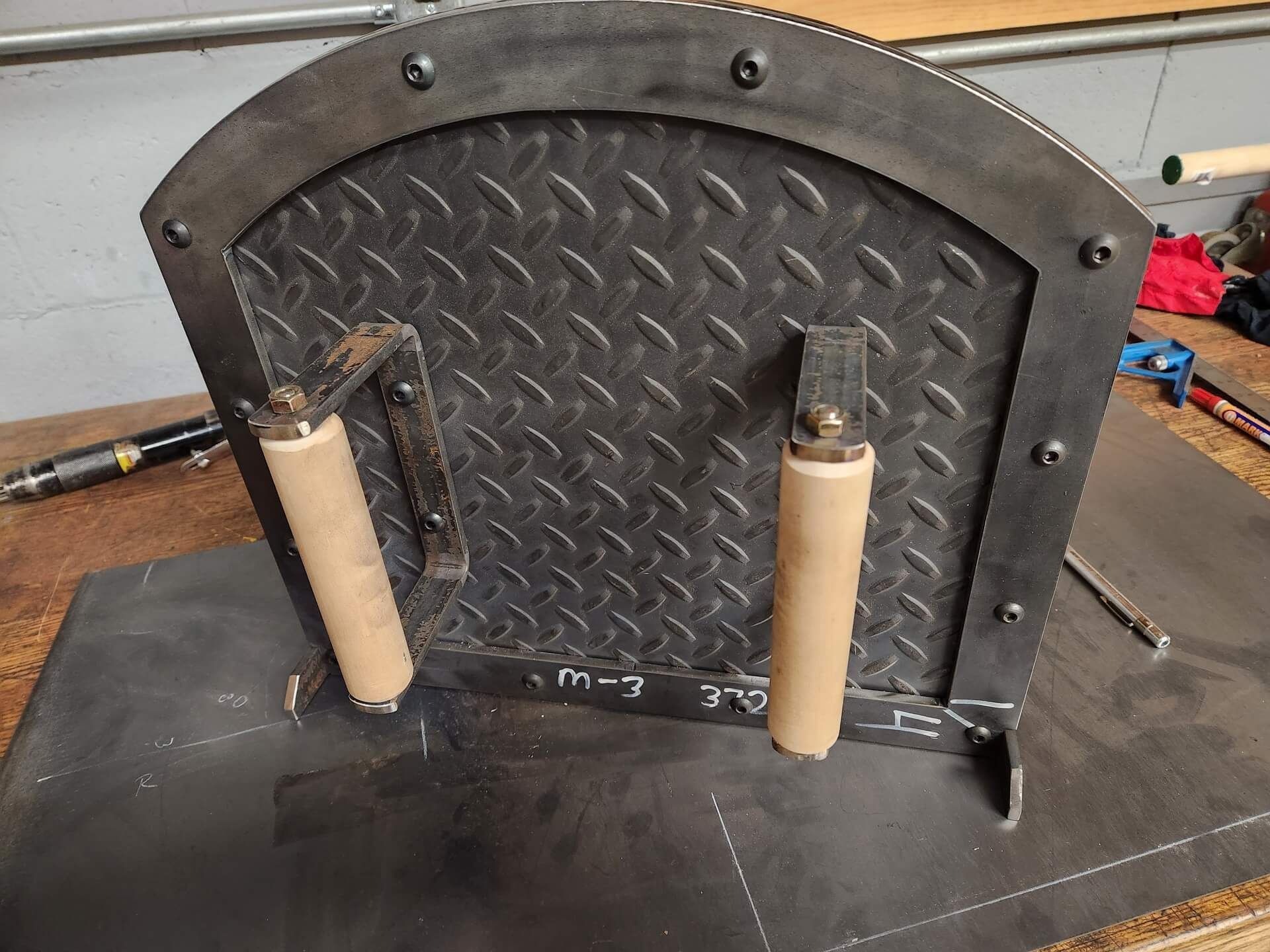 Metal oven door with diamond plate, wooden handles, and rivets. Mounted on a steel base, inside a workshop.