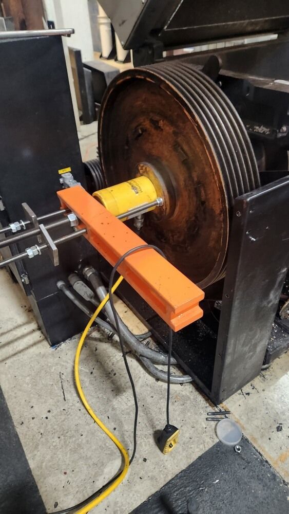Large industrial machine with a rusty wheel, orange safety bar, and yellow cable.