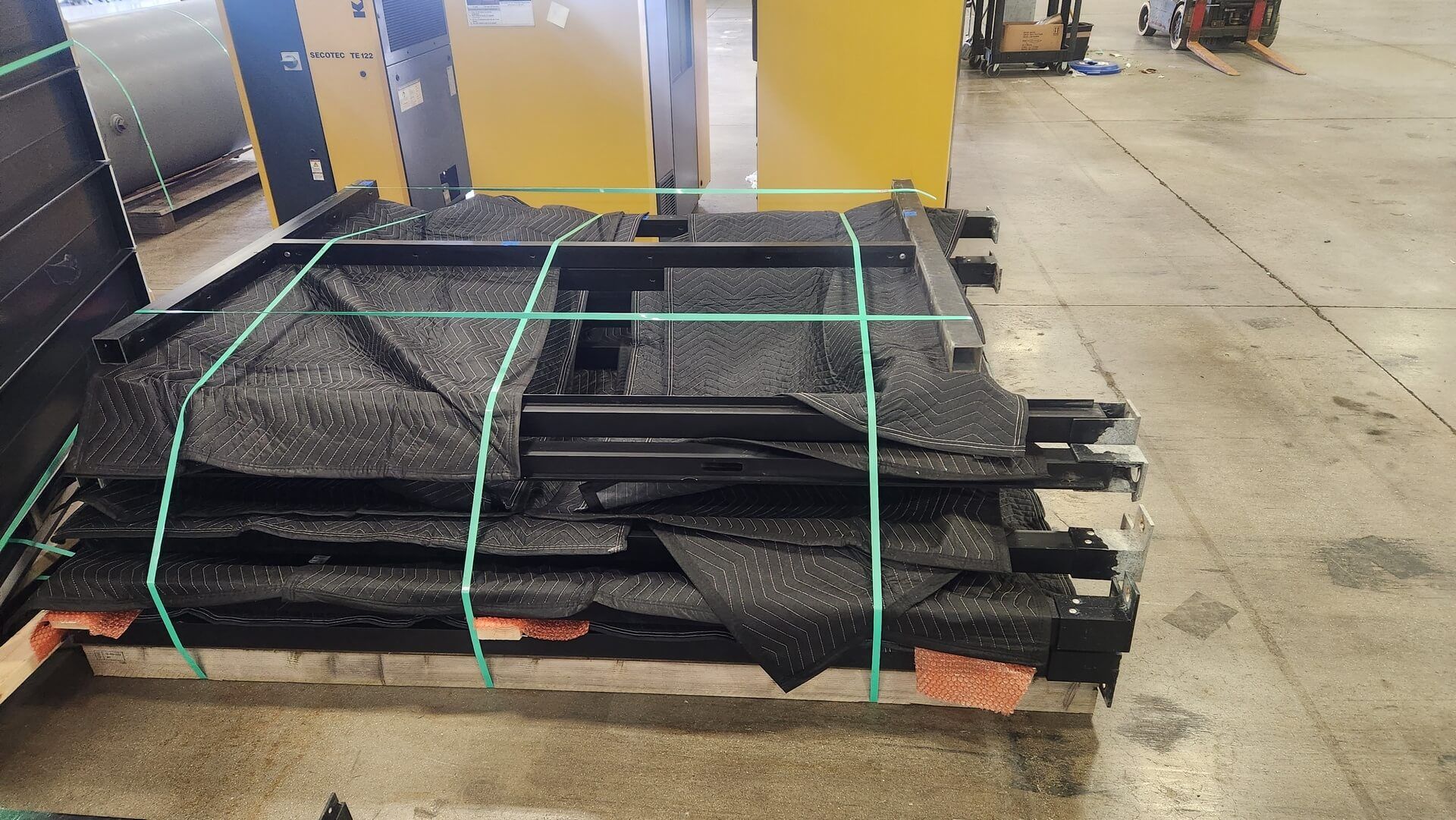 Black metal frames wrapped in black bubble wrap, secured with green straps on a pallet.