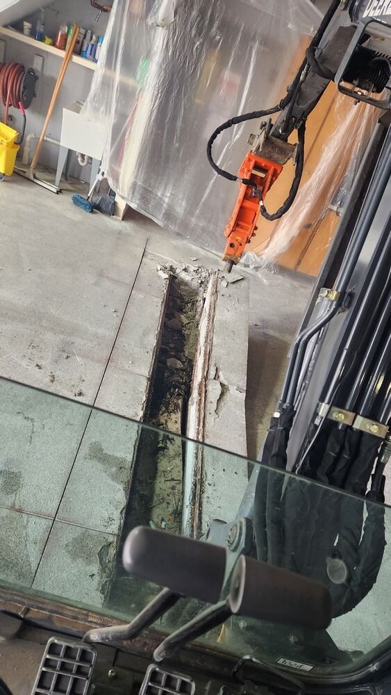 An excavator with a jackhammer breaking up concrete floor inside a building.