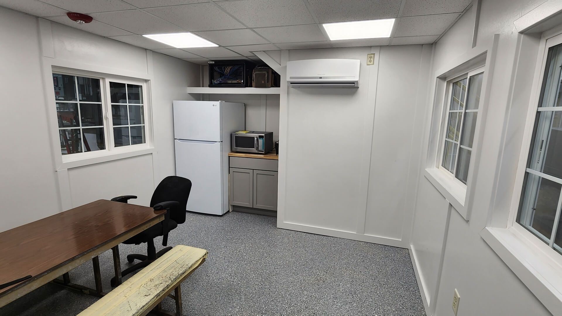 Interior of a small, white-walled room with a table, fridge, microwave, windows, and bench.