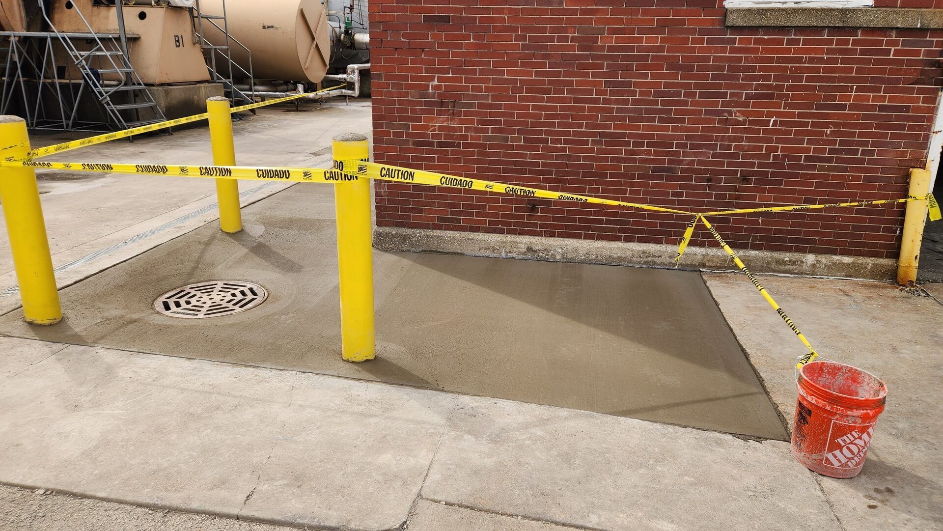 Freshly poured concrete area, bordered by yellow posts and caution tape, next to a red brick building. A red bucket sits on the right.