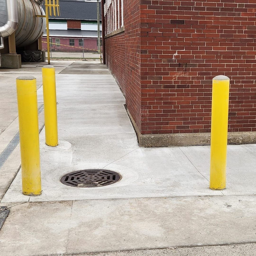 Yellow bollards protect a concrete walkway next to a brick building and a metal tank.