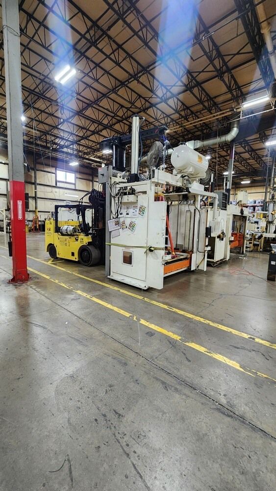 Yellow forklift next to large industrial machinery in a factory. Overhead lighting, yellow floor markings.