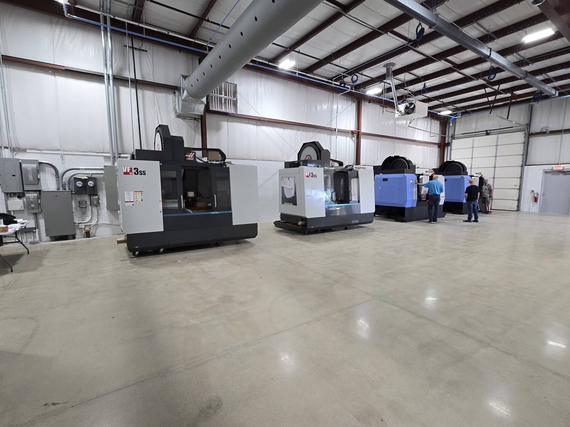 Machining equipment in a warehouse. Several CNC machines are lined up. Two people stand near the equipment.