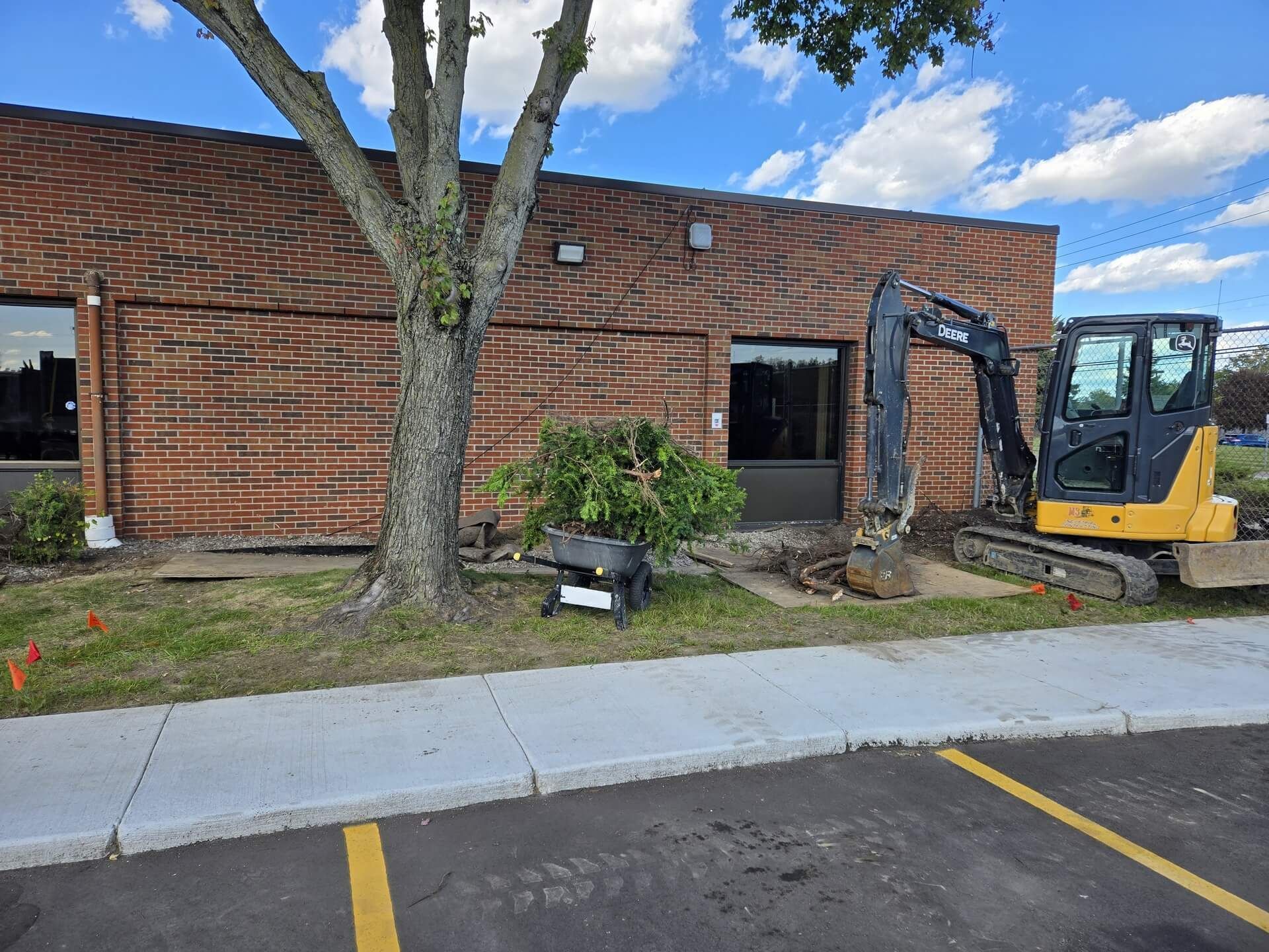 Construction site with backhoe next to a brick building. Trees and grass are visible.