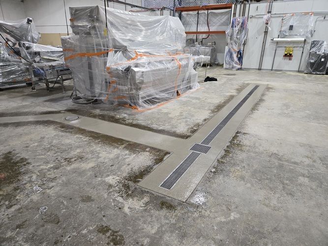 Concrete floor with L-shaped drain, in industrial setting, with wrapped machinery in the background.