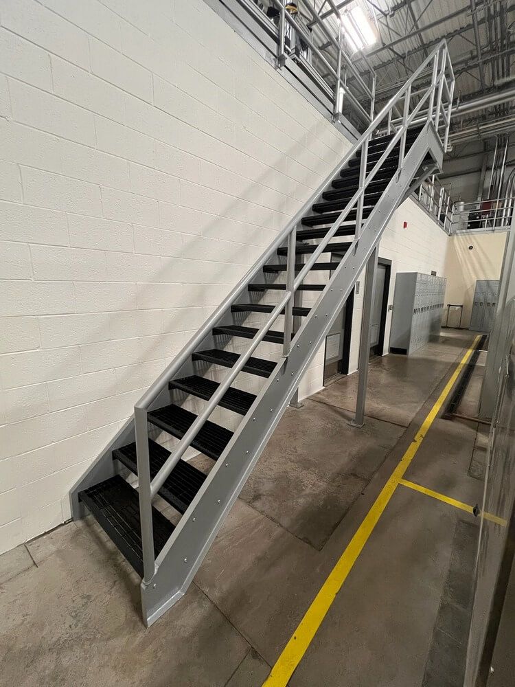 Steel staircase with handrails, leading upwards against a white wall in an industrial setting.