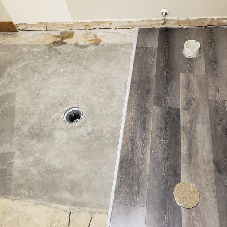 Bathroom floor partially covered with grey wood-look vinyl, concrete floor exposed with drain, new floor installed.