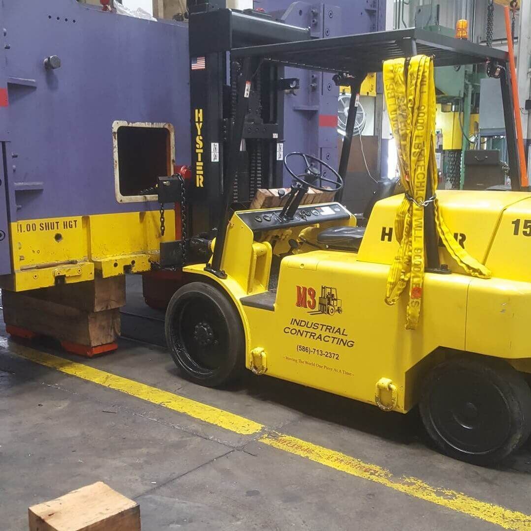 Yellow forklift moving a large, purple industrial machine in a factory.
