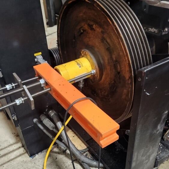 A large industrial wheel with belts and an orange safety fixture, and a yellow cylinder.