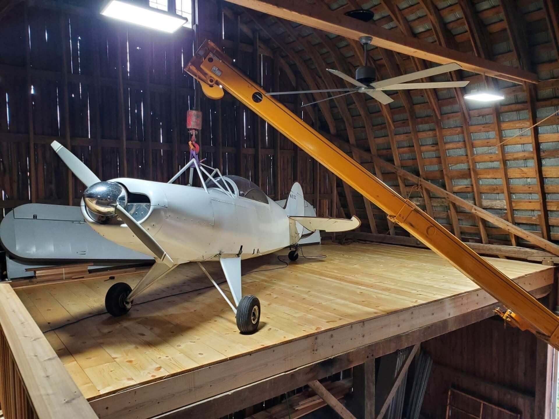 A small, silver airplane inside a wooden barn. The plane is parked on a raised platform, with a beam overhead.