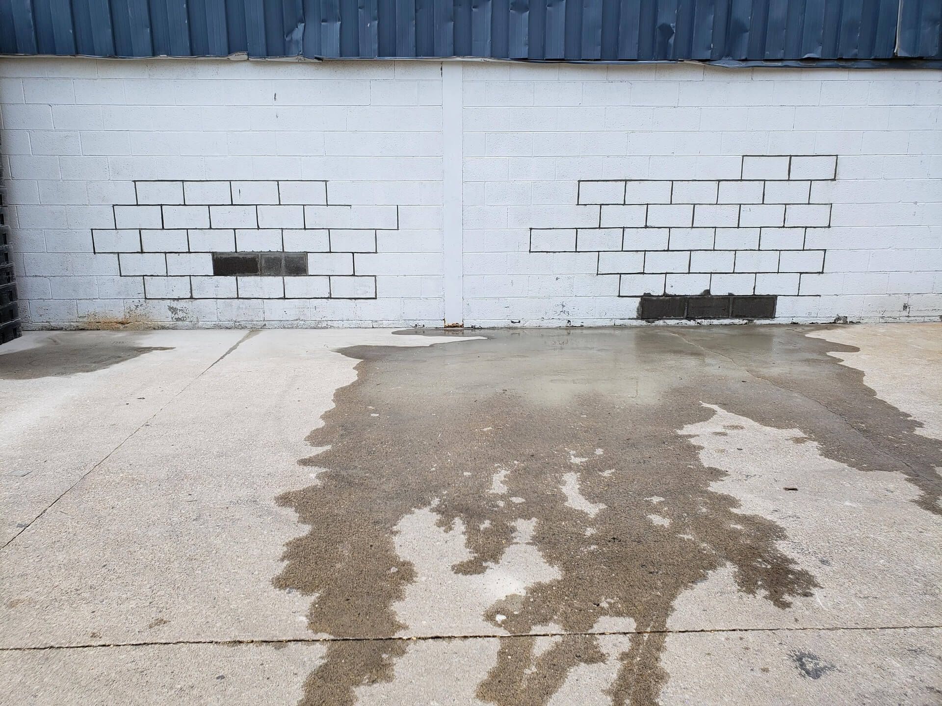 Wet concrete surface with water runoff in front of a white wall with rectangular brick patterns, near a dark blue wooden structure.