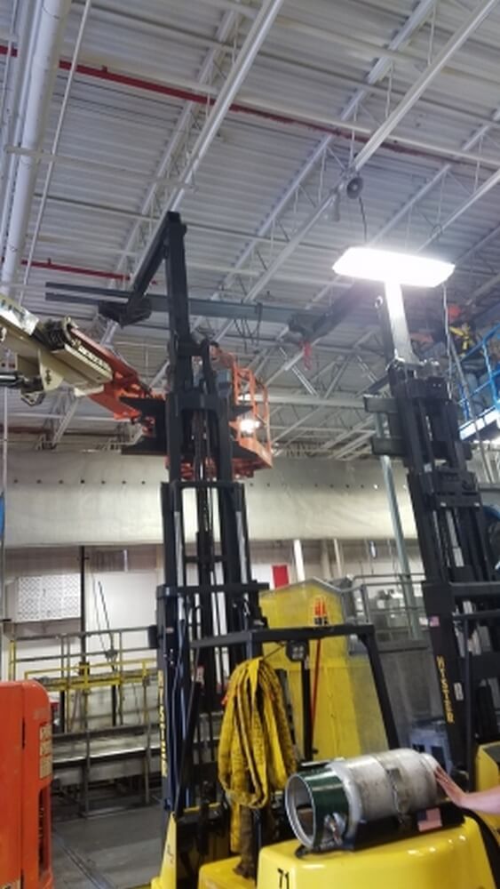 Two forklifts in a warehouse, one lifting a scissor lift. High ceilings with pipes and lights.