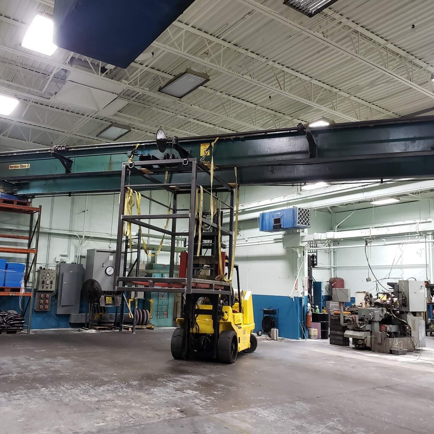 Yellow forklift in a factory lifting a metal frame towards an overhead crane.