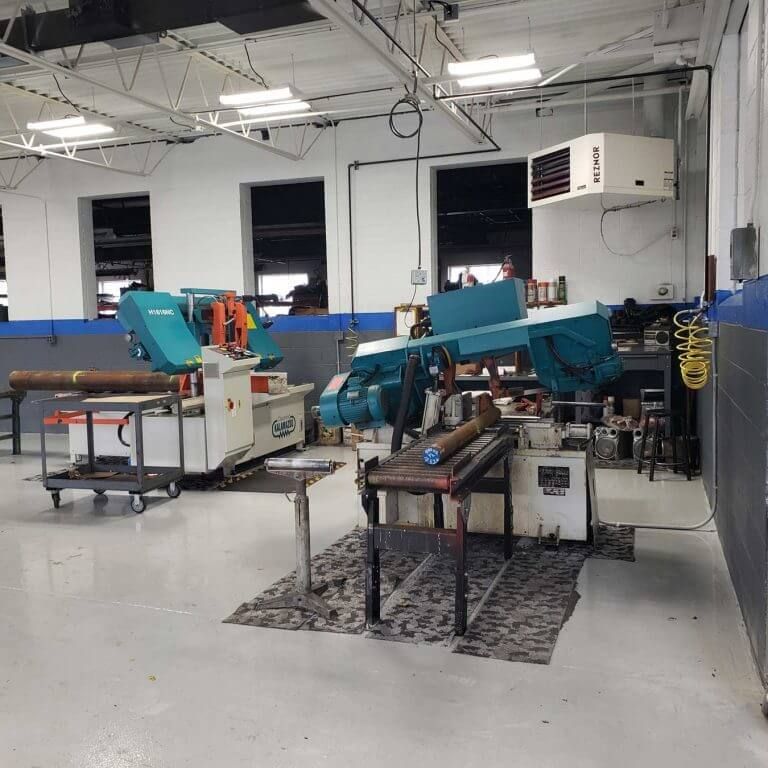 Industrial shop with two large band saws and equipment, a white ceiling and large windows.