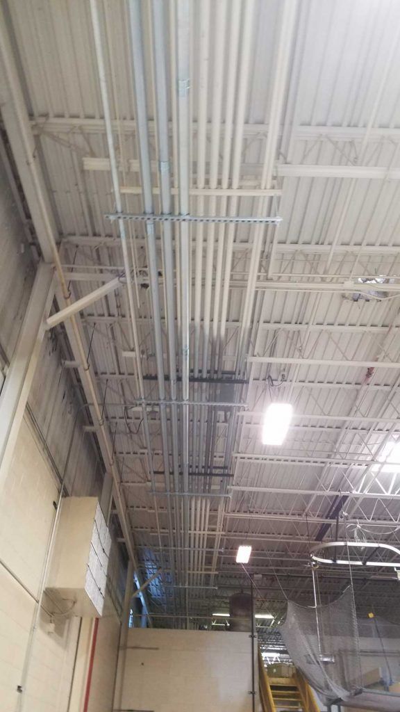 Overhead view of electrical conduit pipes running across a white industrial ceiling, supported by metal brackets.