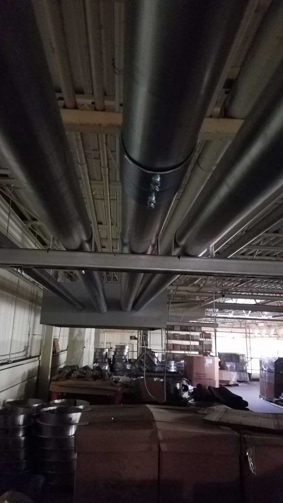 Black HVAC ductwork runs across a warehouse ceiling, with boxes and equipment visible below.