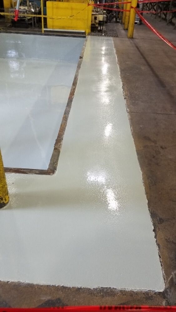 A newly painted industrial floor with light gray sections and unfinished edges, safety tape in foreground.
