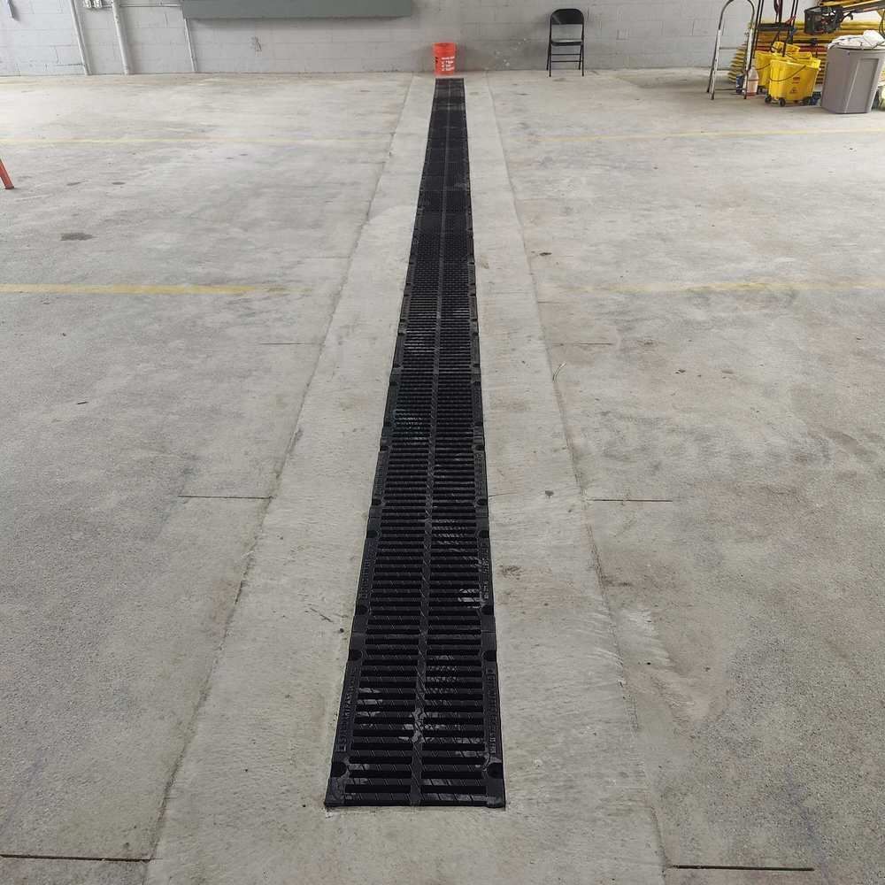 A long, black drain grate in a concrete floor, likely in a garage or industrial setting.