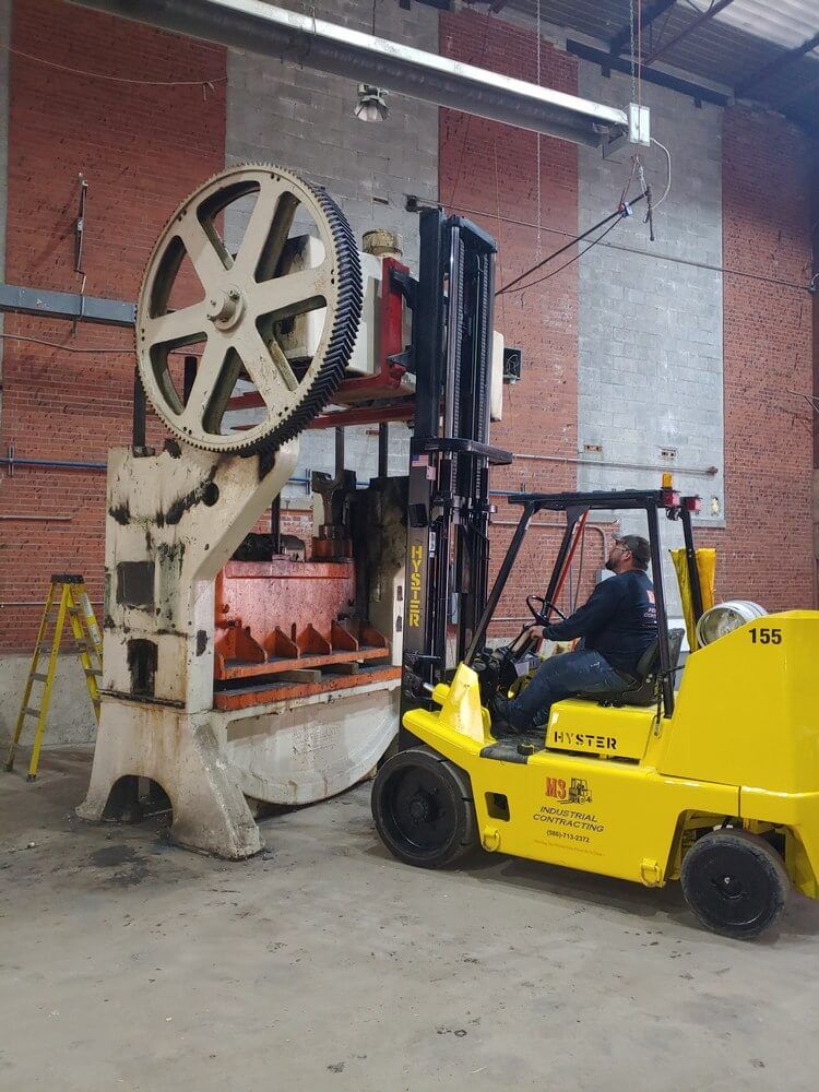 Forklift lifting machinery in a brick building. Operator in cab, large gear wheel visible.
