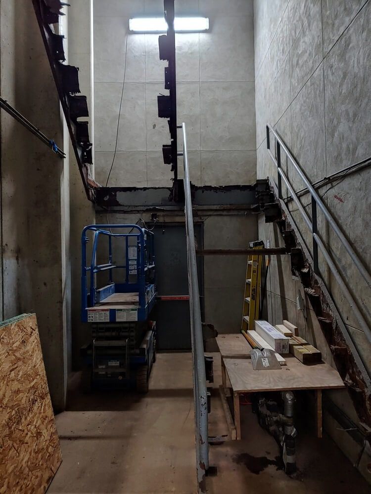 Interior view with stairs, a lift, and a workbench. Industrial setting with concrete walls and overhead lighting.