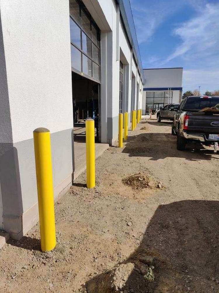 Yellow bollards protect a building's entrance. Dirt ground. Trucks parked near open garage doors. Blue sky.
