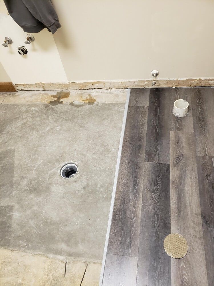 Bathroom floor: concrete subfloor next to installed wood-look vinyl plank flooring. Drain visible.