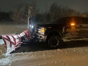 Snowplow clearing snow from a driveway at night. Dark truck with a red and white plow, with snow-covered setting.