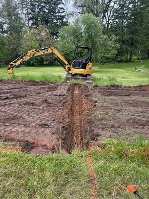 Yellow excavator digging trench in muddy field, orange lines mark boundaries, trees in background.