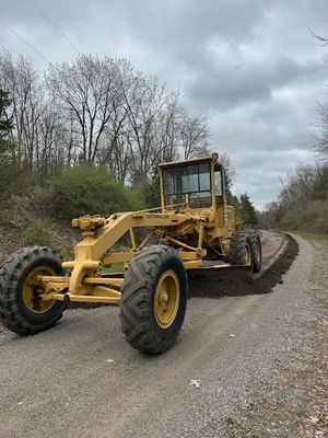 Yellow grader on a gravel road, grading the side. Cloudy sky, trees in background.