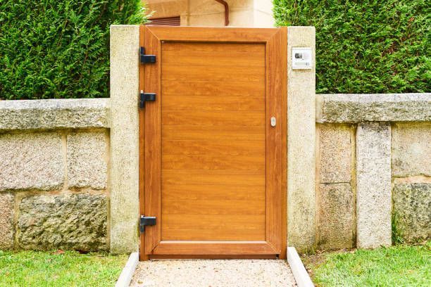 Wooden gate, brown frame, black hinges, set in stone wall, green hedge background.