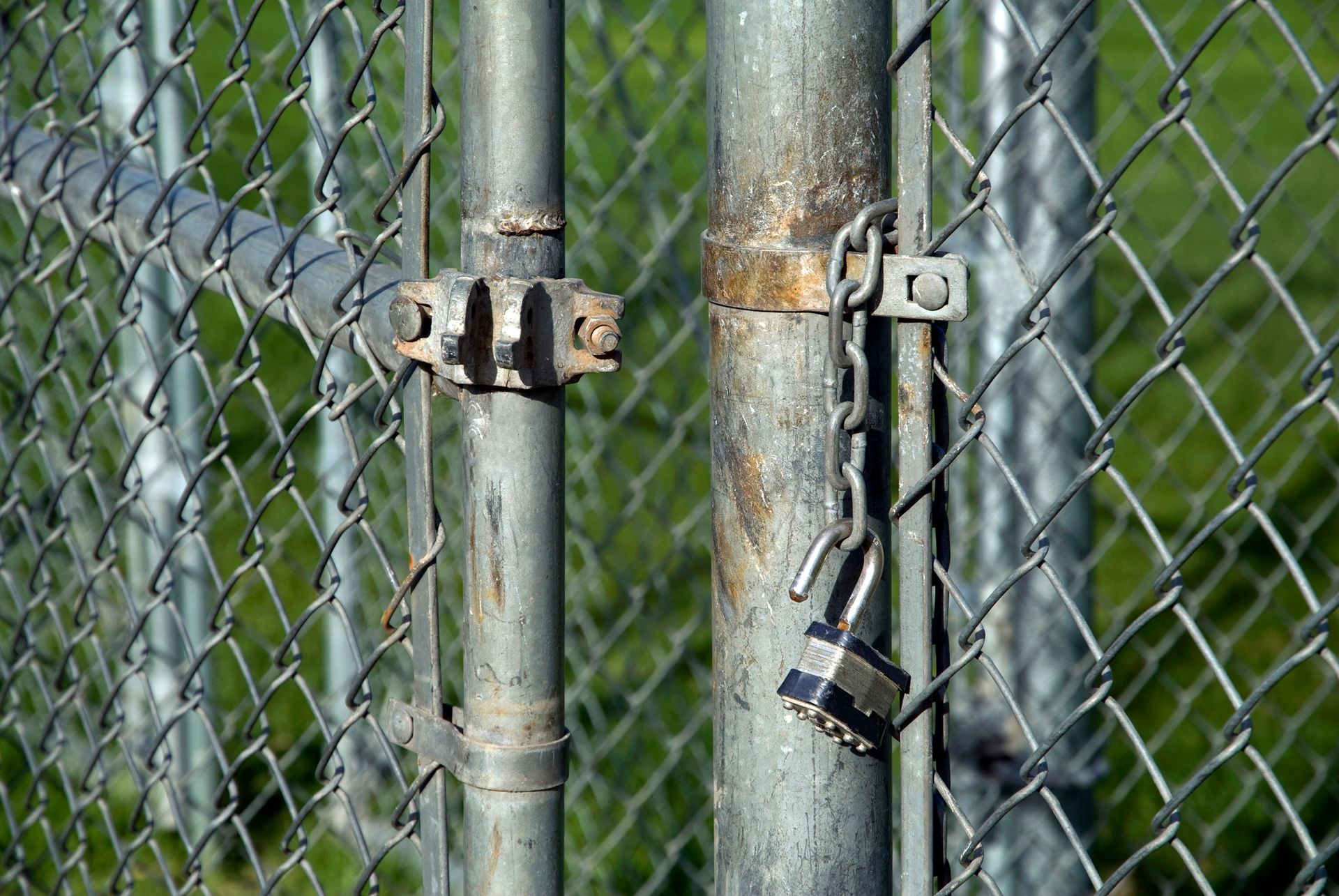 Locked chain-link fence gate. Silver metal gate posts with a padlock. Green grass background.