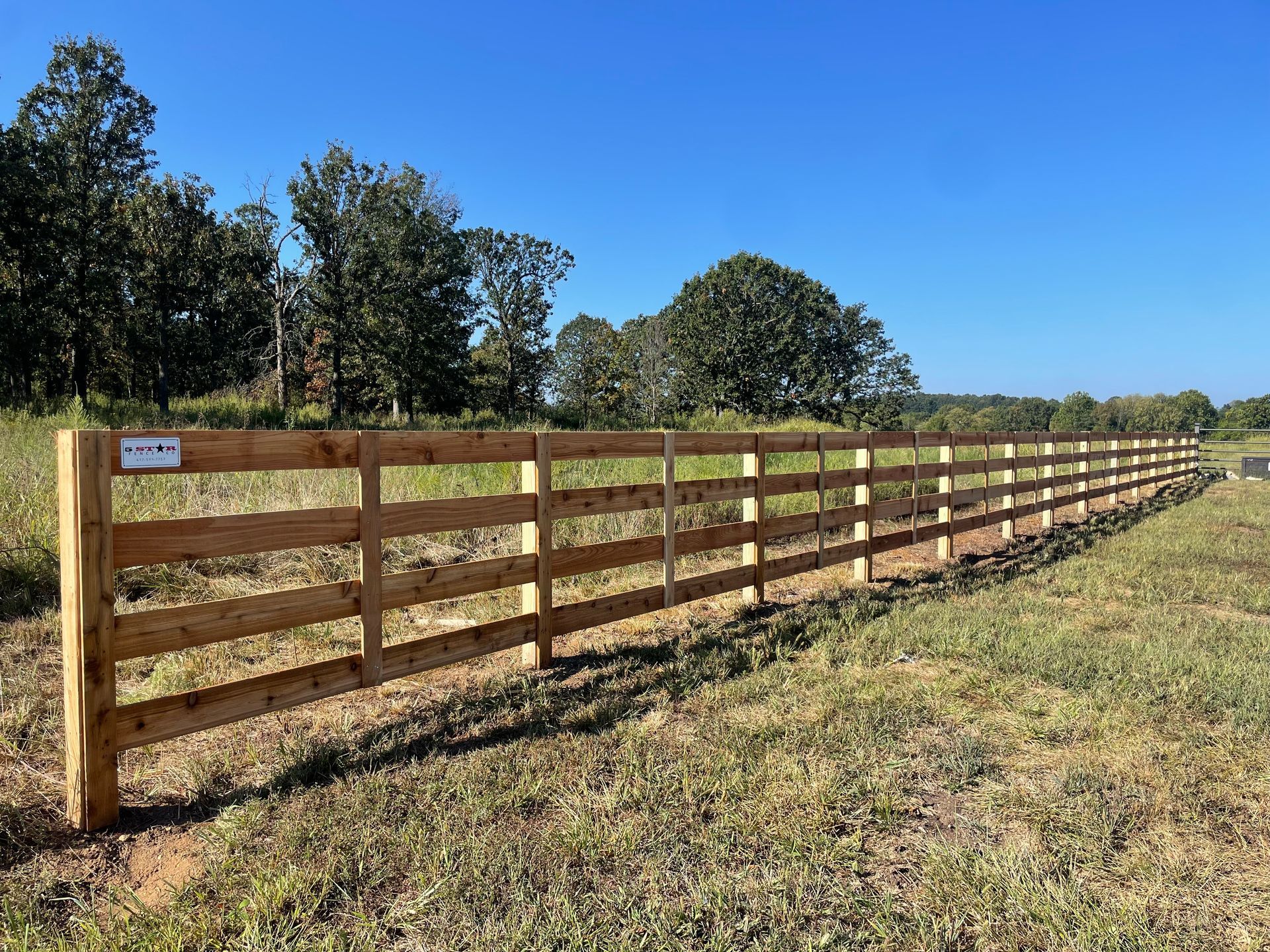Brown wooden fence alongside a concrete walkway with grass and trees in the background.
