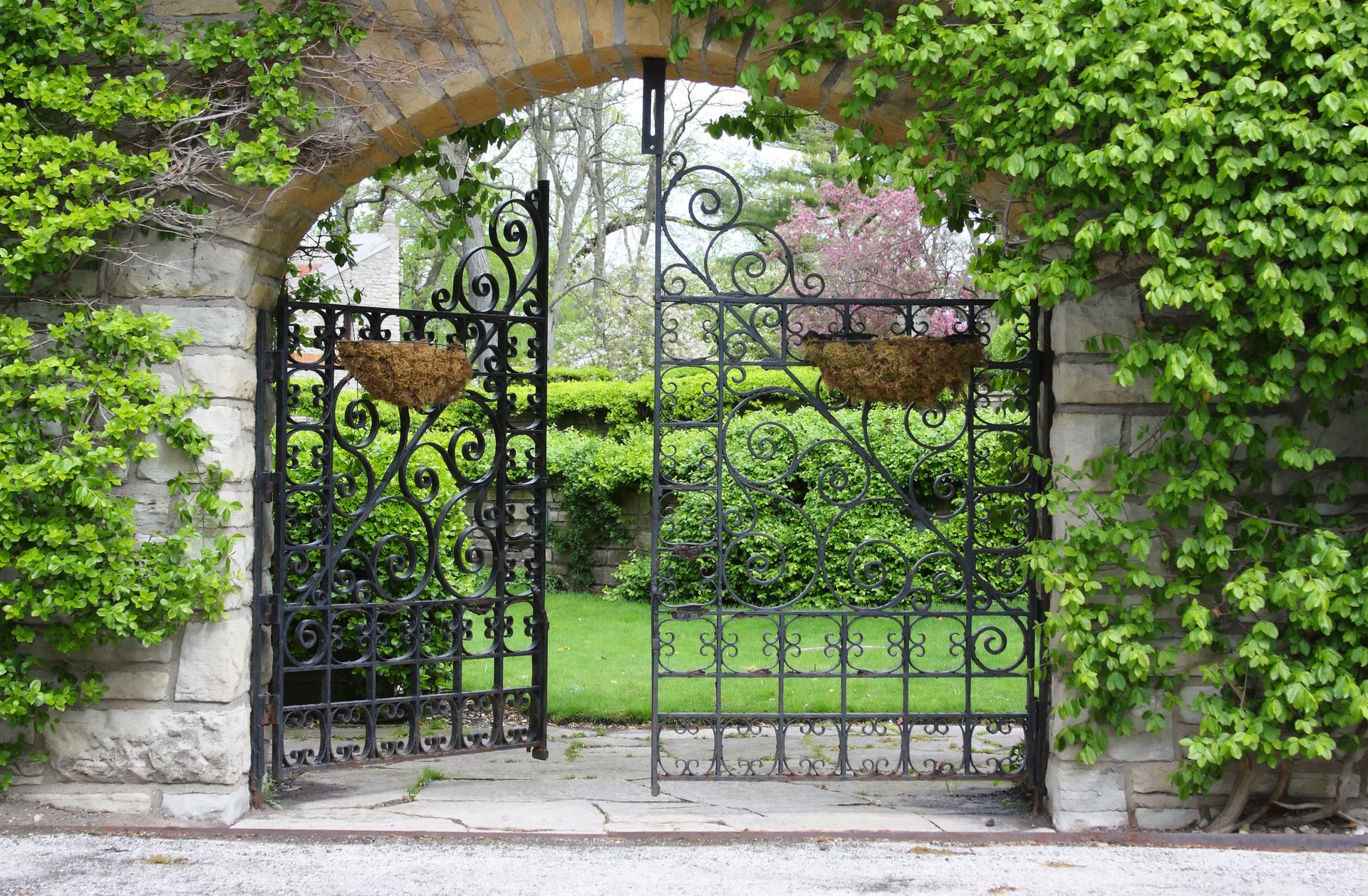 Ornate black gate opening into a garden, framed by a stone arch covered in green foliage.