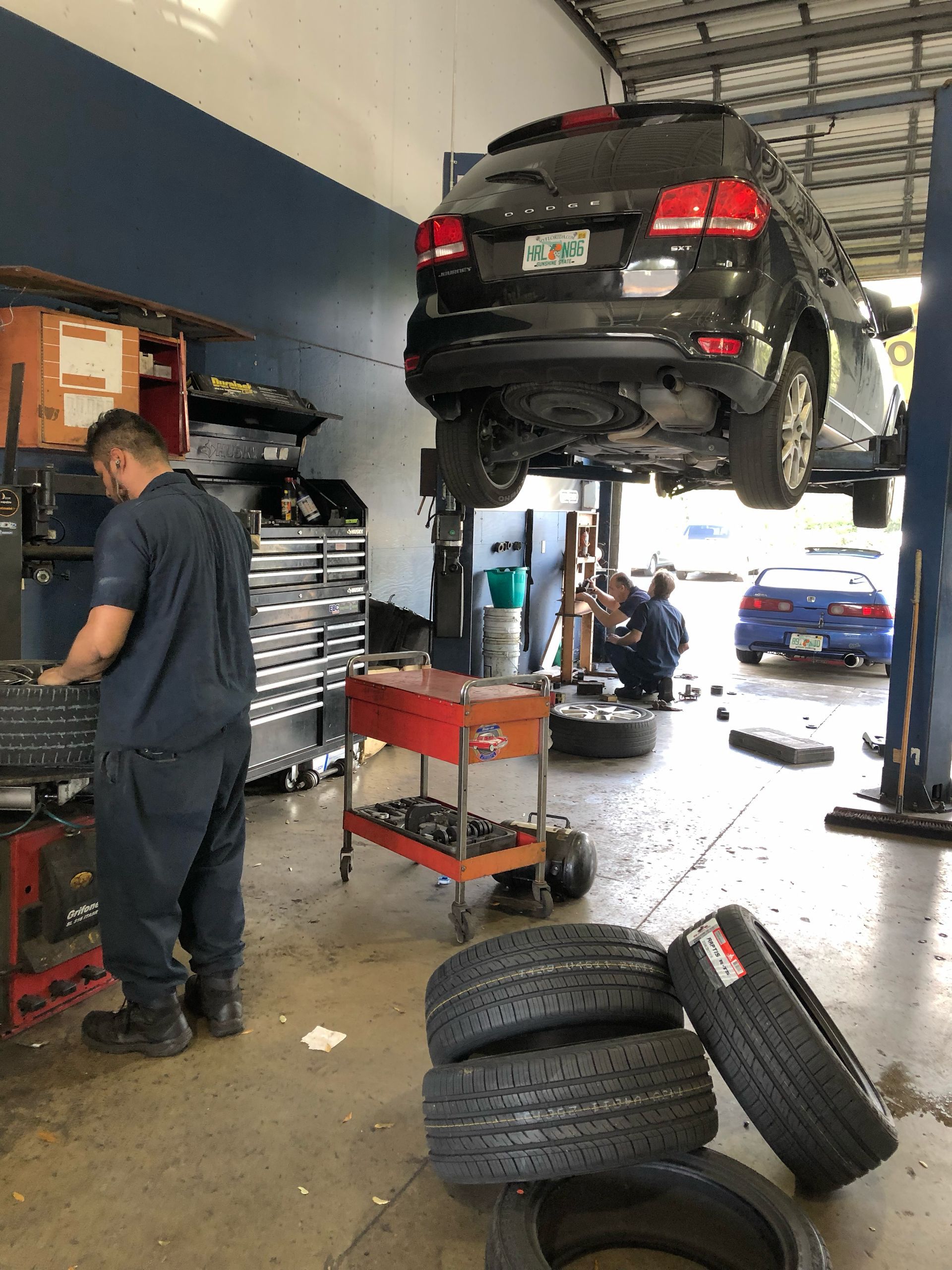 Mechanics working in a garage. A black SUV is lifted. Tires stacked on the floor. One mechanic at a toolbox. | Brake World