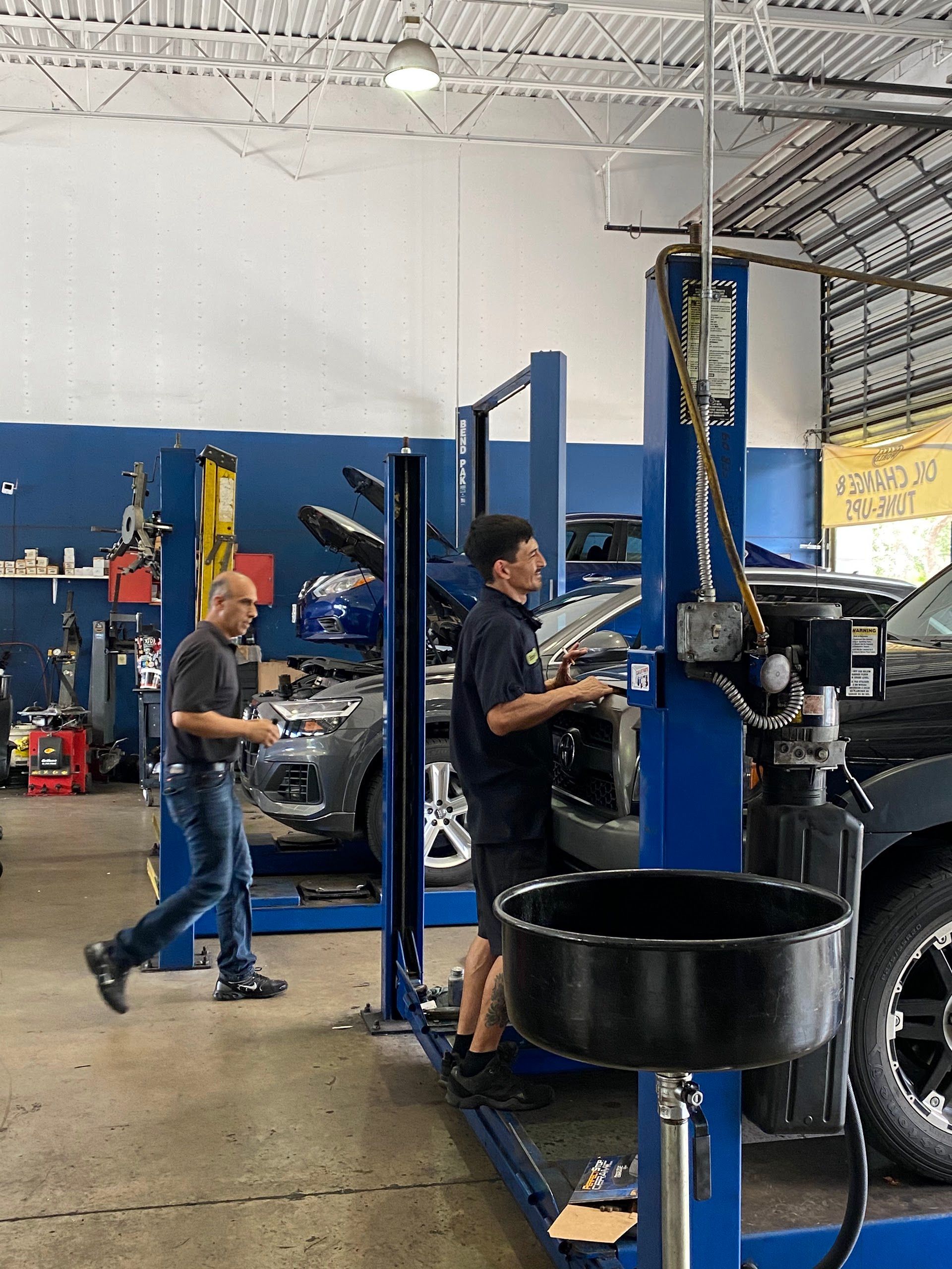 Two mechanics working in an auto repair shop. One adjusts a lift, the other walks past a car. Blue and white walls. | Brake World