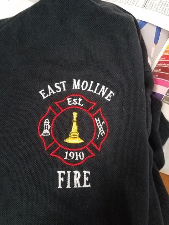 Embroidered logo of the East Moline Fire Department on a black shirt. The logo features a red Maltese cross with the city name, 