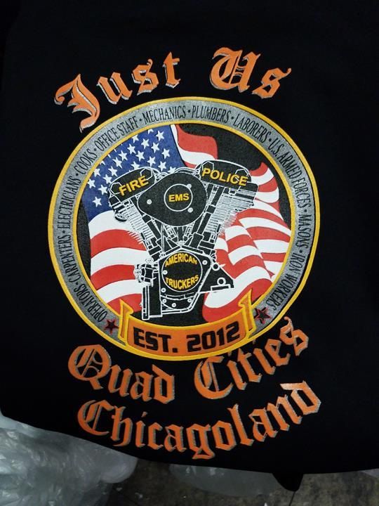 Black shirt with a circular logo featuring an American flag, motorcycle engine, and text for “Just Us Quad Cities Chicagoland.”