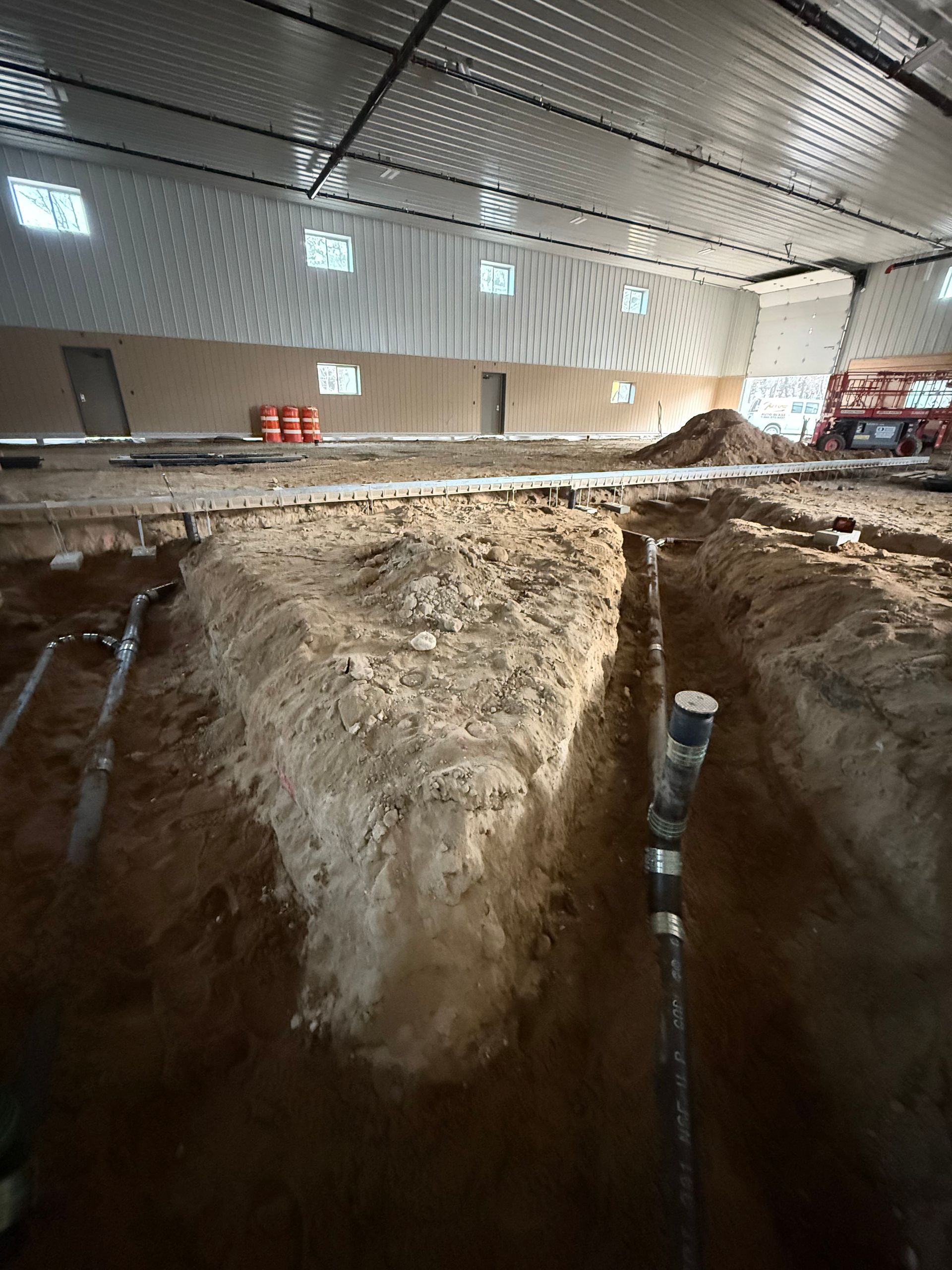 Interior of a building under construction, with trenches dug in the dirt floor. Pipes and equipment are visible.