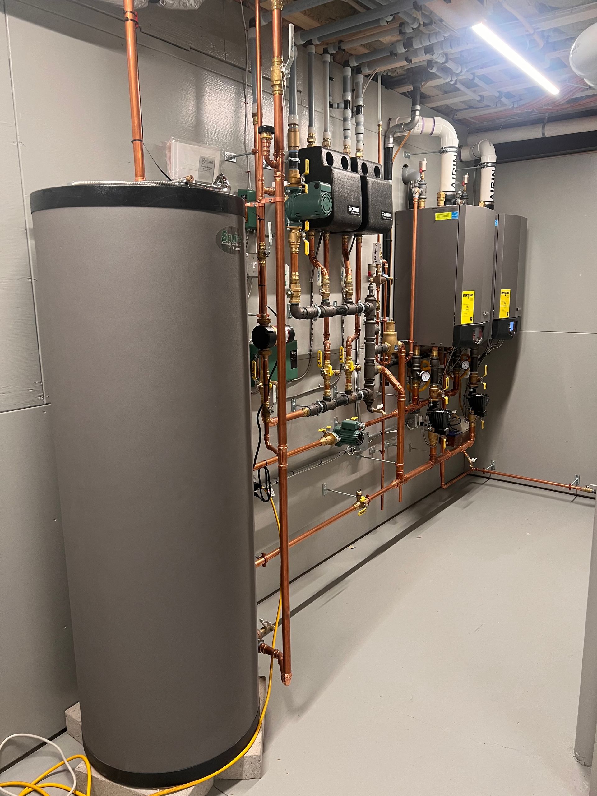 Gray and copper plumbing system in a utility room; a large water tank, pipes, and control panels.