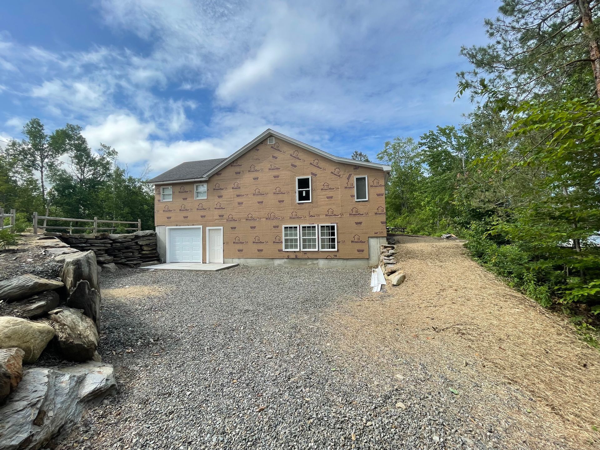 Two-story house with gravel driveway and rock retaining wall, surrounded by trees under a cloudy sky.