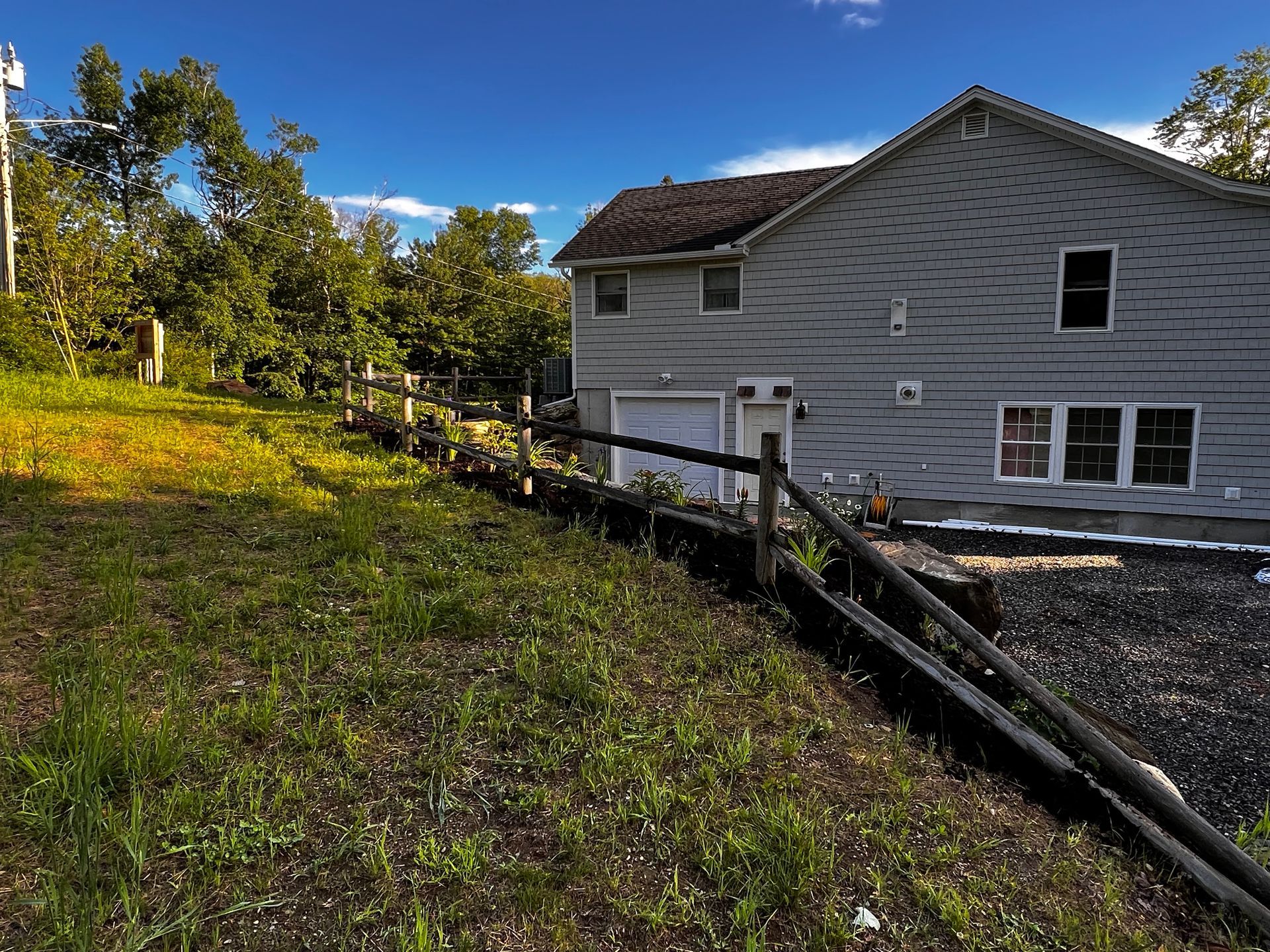 A gray two-story house with a garage, seen from a sloped grassy yard with a wooden retaining wall.