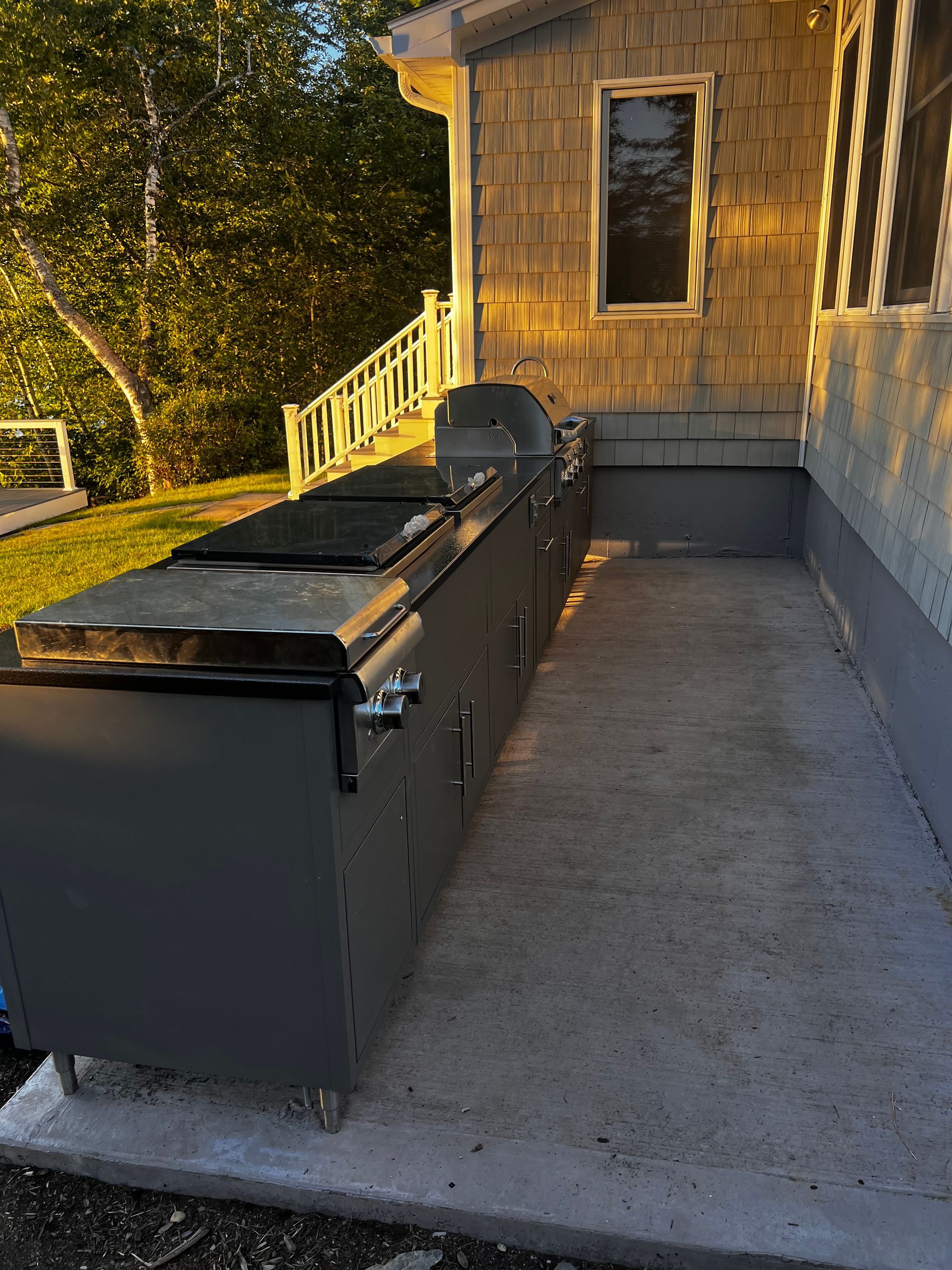 Outdoor kitchen with stainless steel appliances and concrete patio. Building with wooden shingles in background.