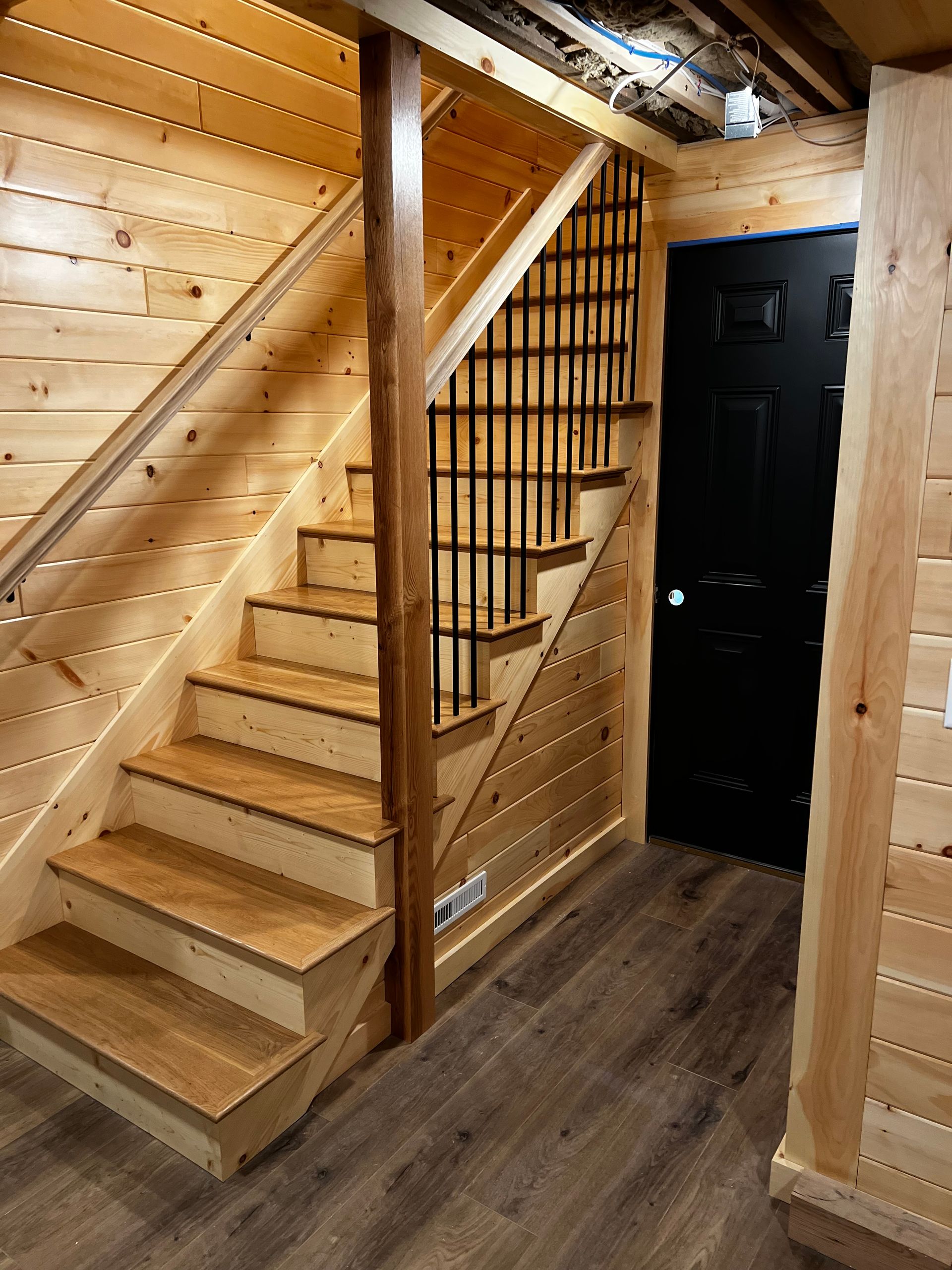 Wooden staircase in a basement, leading up. Black door on right, brown floor, and tan wood walls.