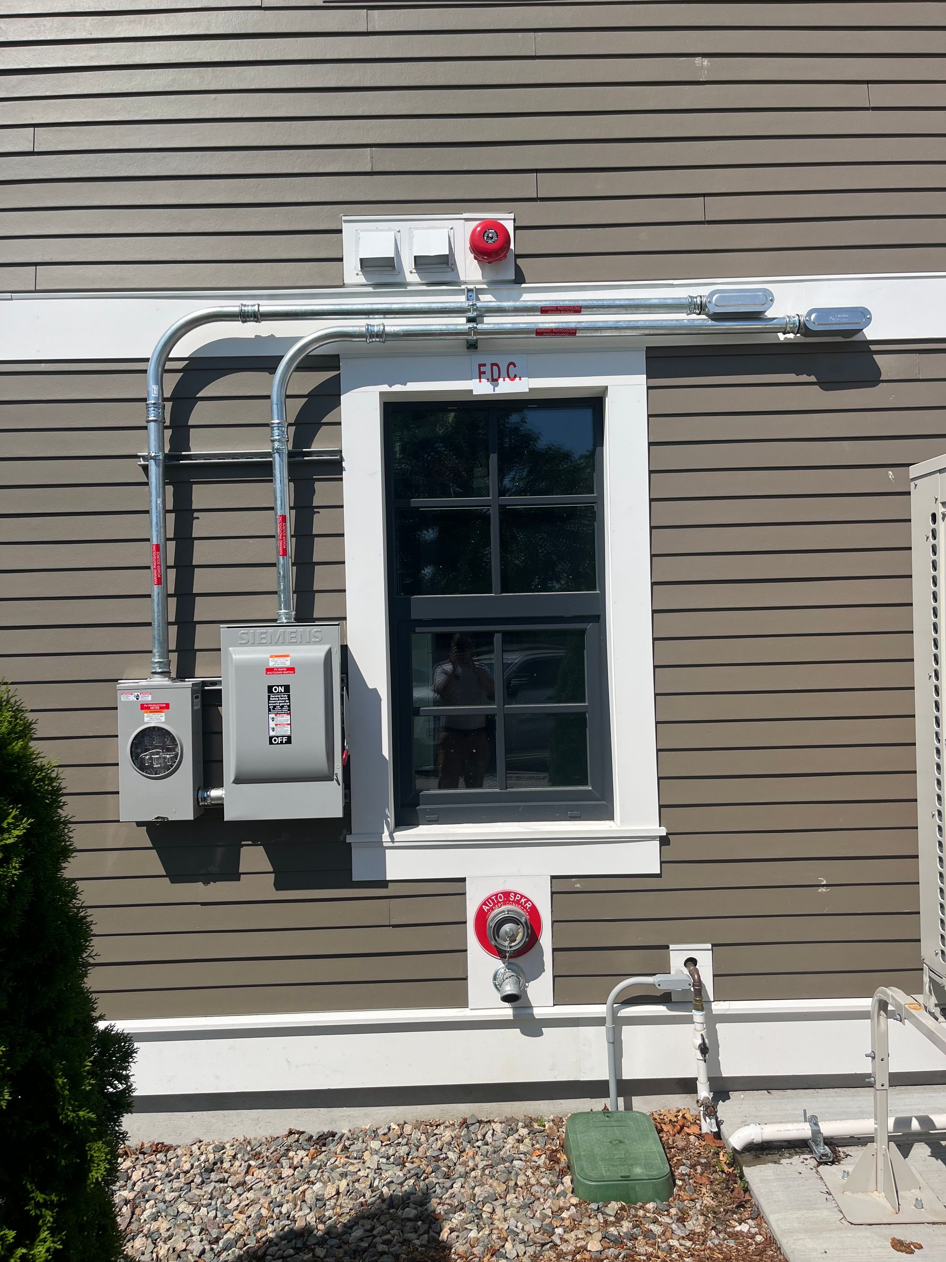 Exterior of a building with electrical equipment and a window. Gray siding, metal conduit, and gray electrical boxes.